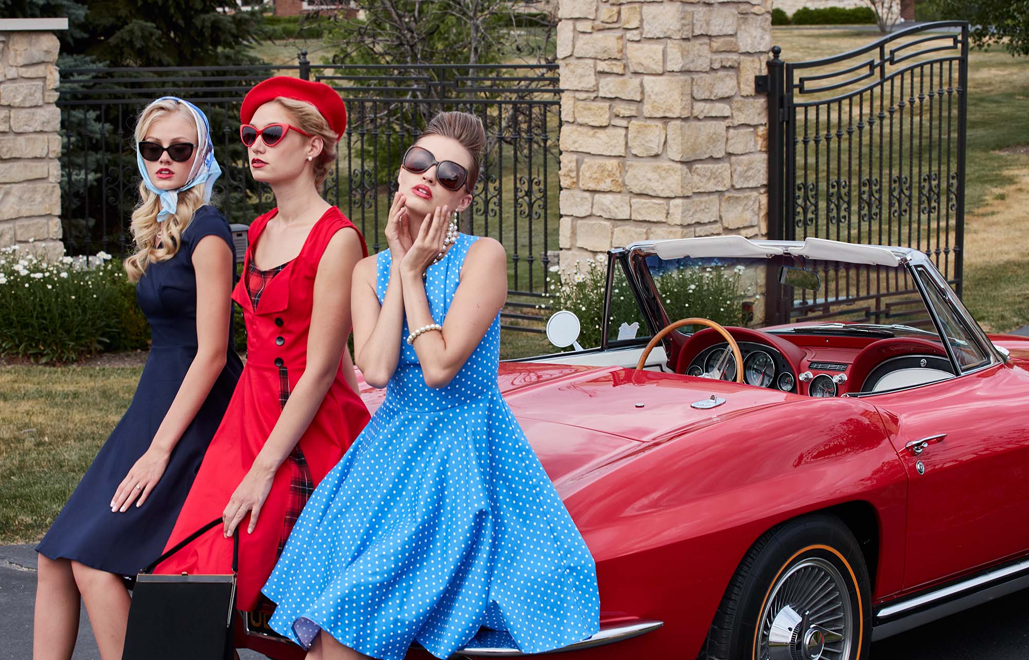 Three women in vintage fashion pose by a red convertible car. They are wearing colorful dresses and retro sunglasses with mid-century hairstyles.
