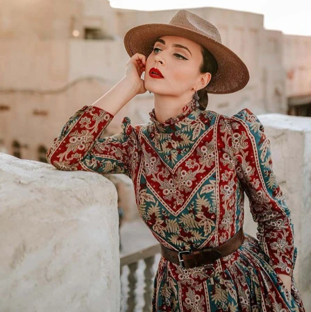 Adult female in a patterned long-sleeve dress and straw hat poses outdoors with hand on her face. Building exterior in background.