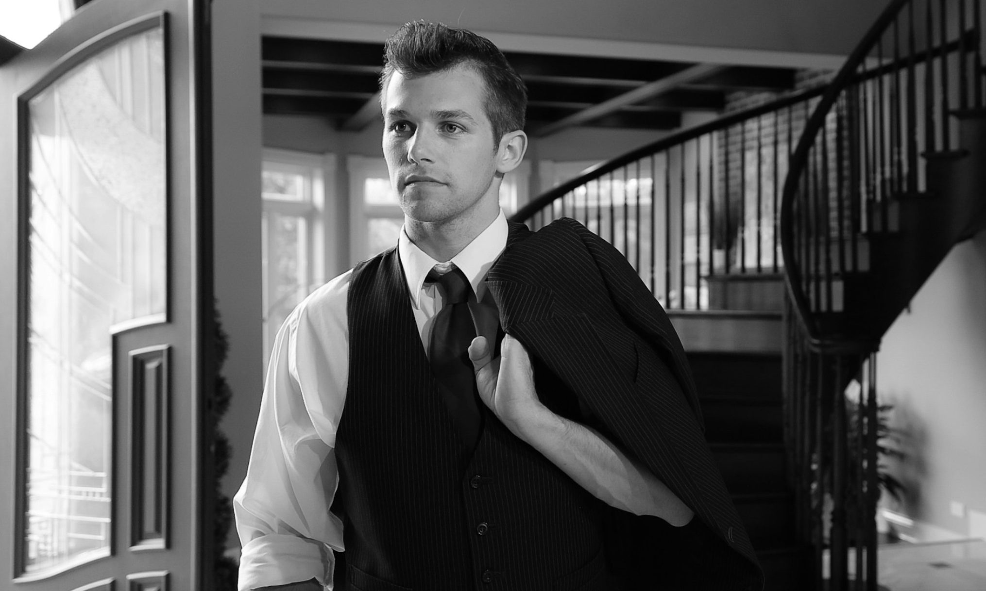 Black and white photo of a man in a pinstripe vest and shirt holding his suit jacket over his shoulder indoors near a staircase.