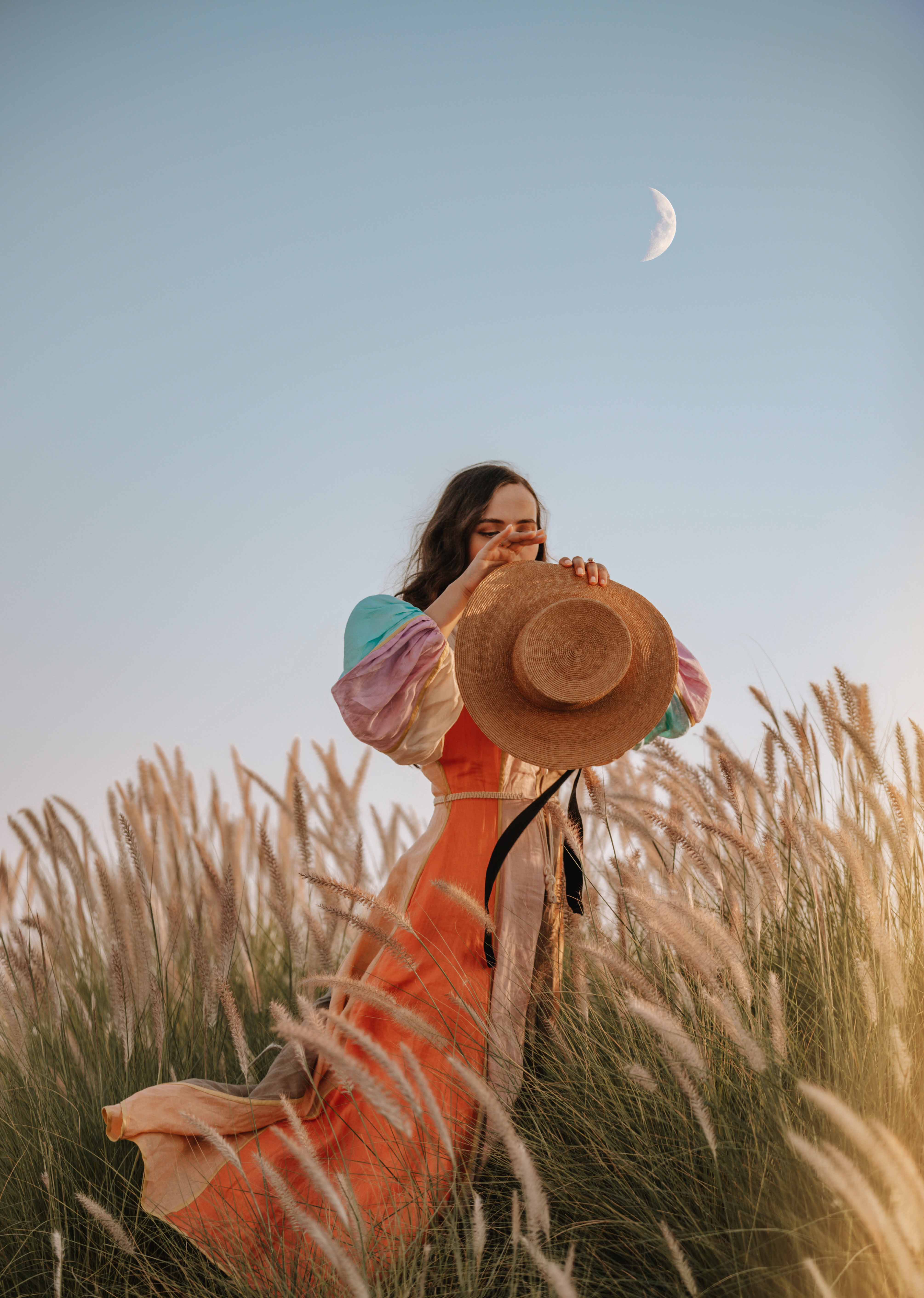 A woman in a colorful dress holds a hat standing in a field of tall grass under a pale blue sky with a crescent moon in the background.