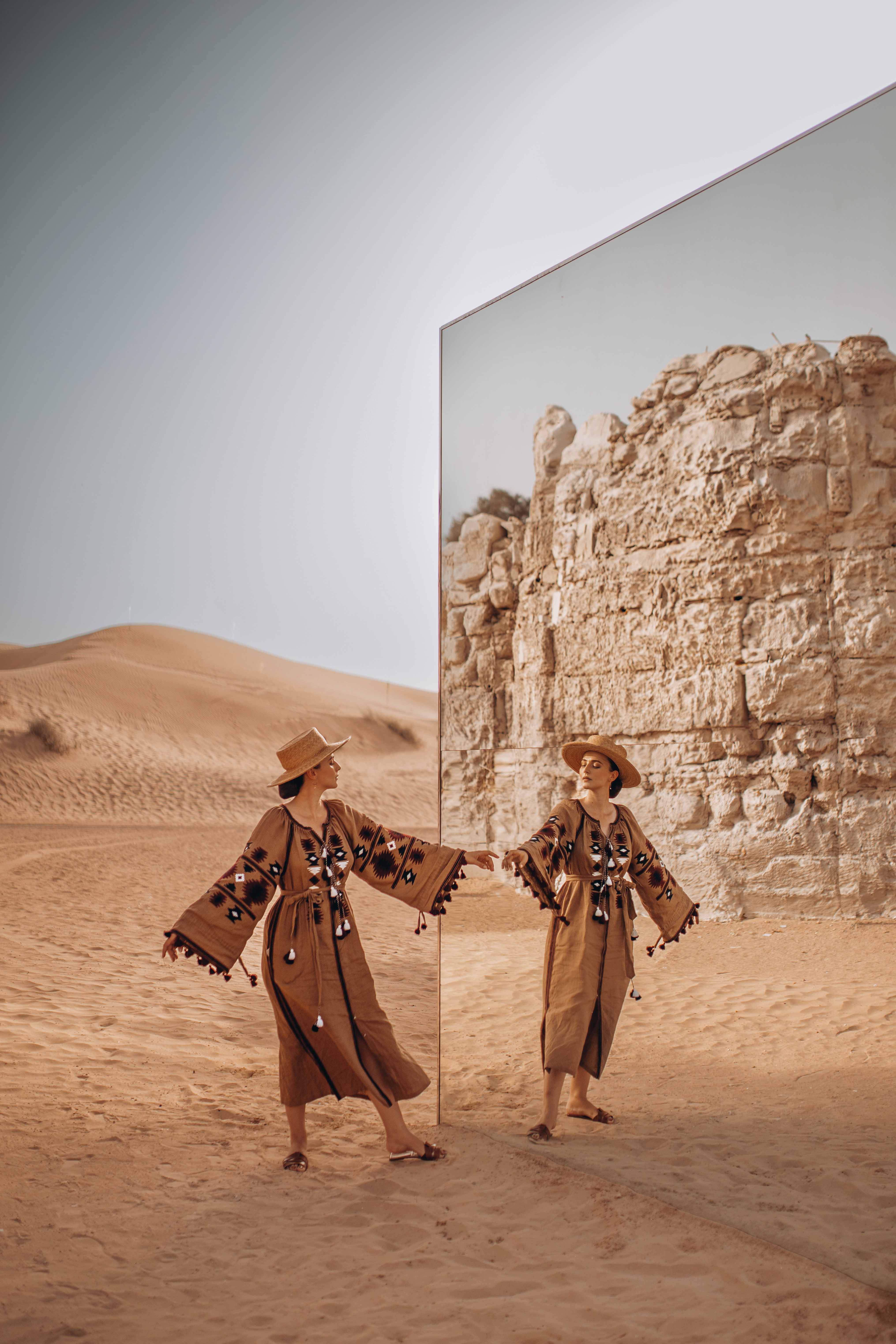 Woman in a tan dress and straw hat poses in a desert near a mirror reflecting a rock wall on a sunny day.  