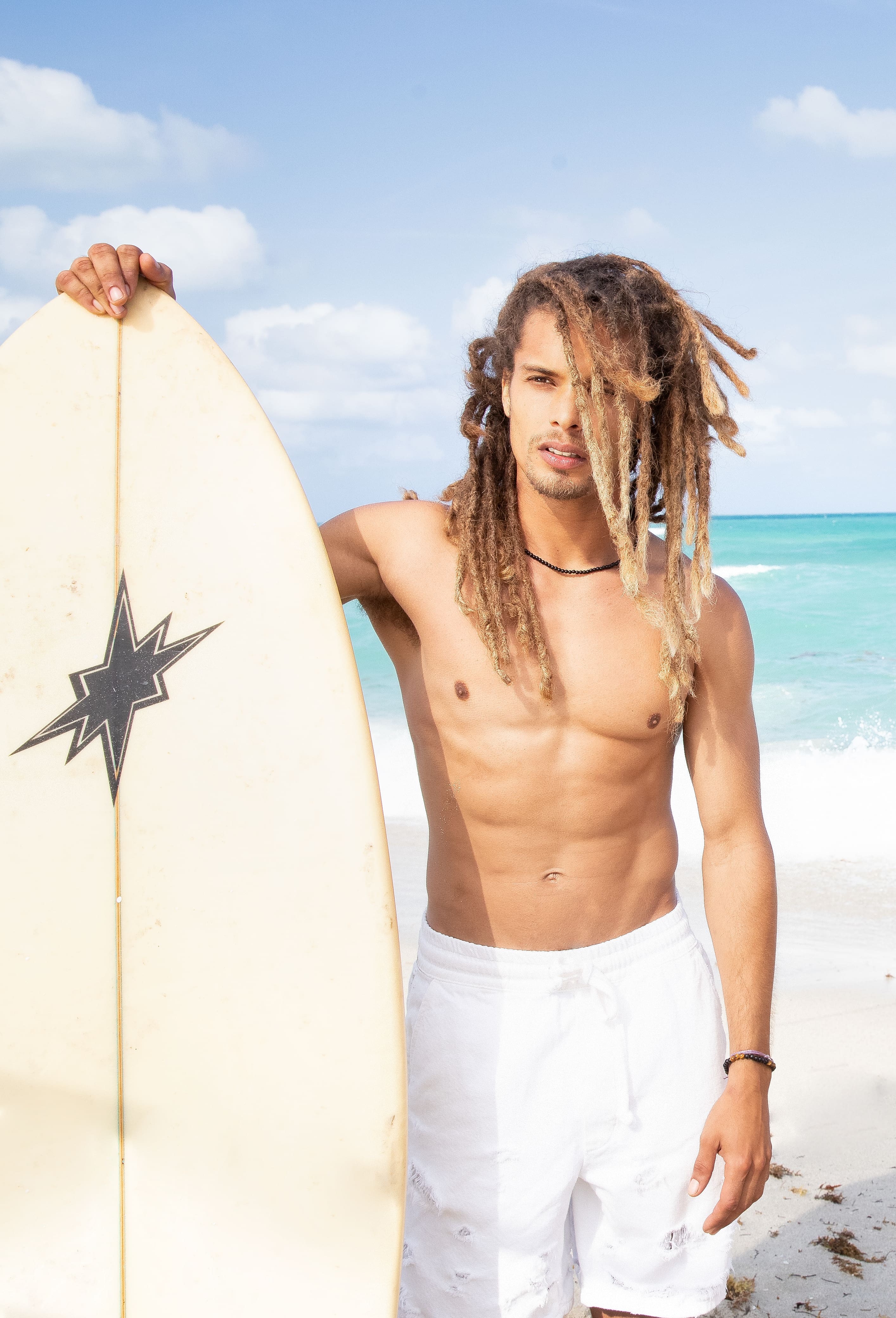 A shirtless man with dreadlocks stands on a white sand beach holding a surfboard with a black star on a sunny day.