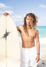 Shirtless man with long dreadlocks holding a surfboard standing on a sandy beach