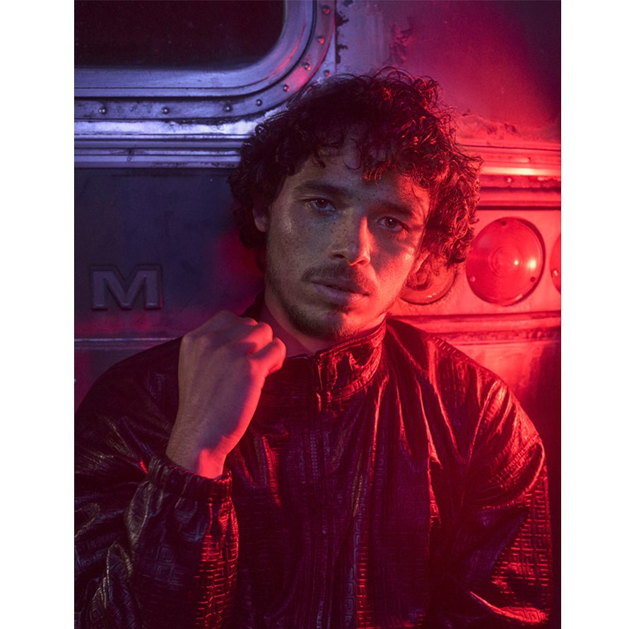 Portrait of a young adult man in a black jacket against a metal background with red and blue lighting in a medium shot.