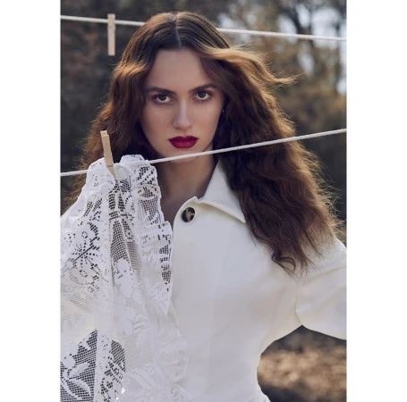 Woman with long brown wavy hair poses in front of a clothesline holding a white lace cloth, wearing a white jacket, portrait shot.