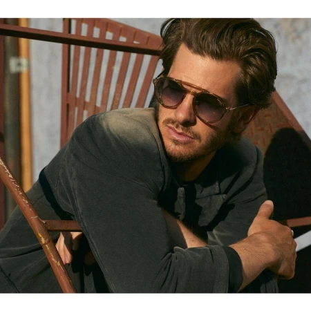 Andrew Garfield wearing gradient aviator sunglasses and a dark jacket outdoors against a rusted metal structure.