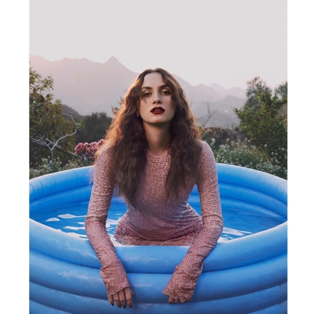 Woman in a pink sequin dress sits in an inflatable kiddie pool in a backyard with mountains in the background at dusk.