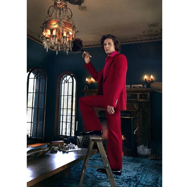 Man in a red suit stands on a ladder dusting an ornate chandelier in a room with dark walls and a fireplace.
