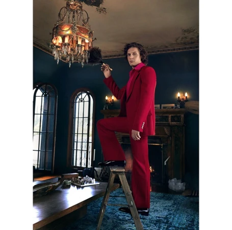 Man in a red suit stands on a ladder dusting an ornate chandelier in a room with dark walls and a fireplace.