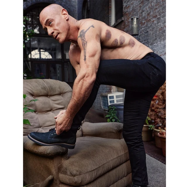 Bald adult male ties black lace-up shoe while leaning on a beige sofa outdoors with a brick wall in the background, artistic tattoo visible.