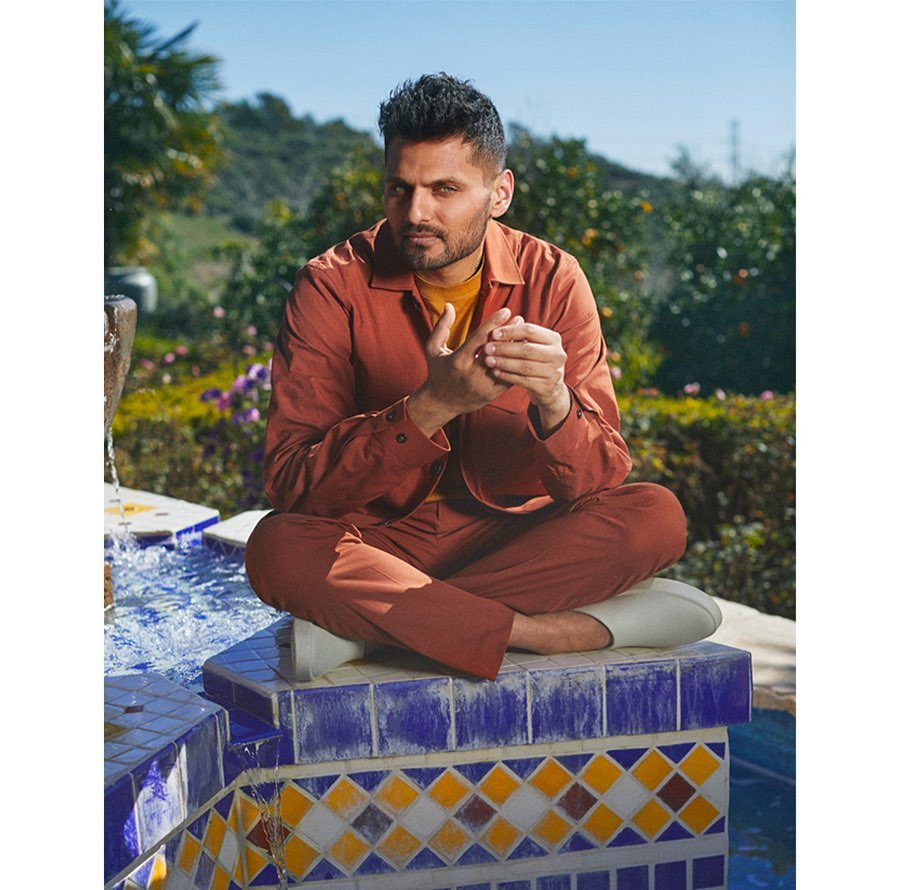 A man in a russet-colored outfit sits cross-legged on a mosaic fountain in an outdoor setting, surrounded by greenery under a blue sky.