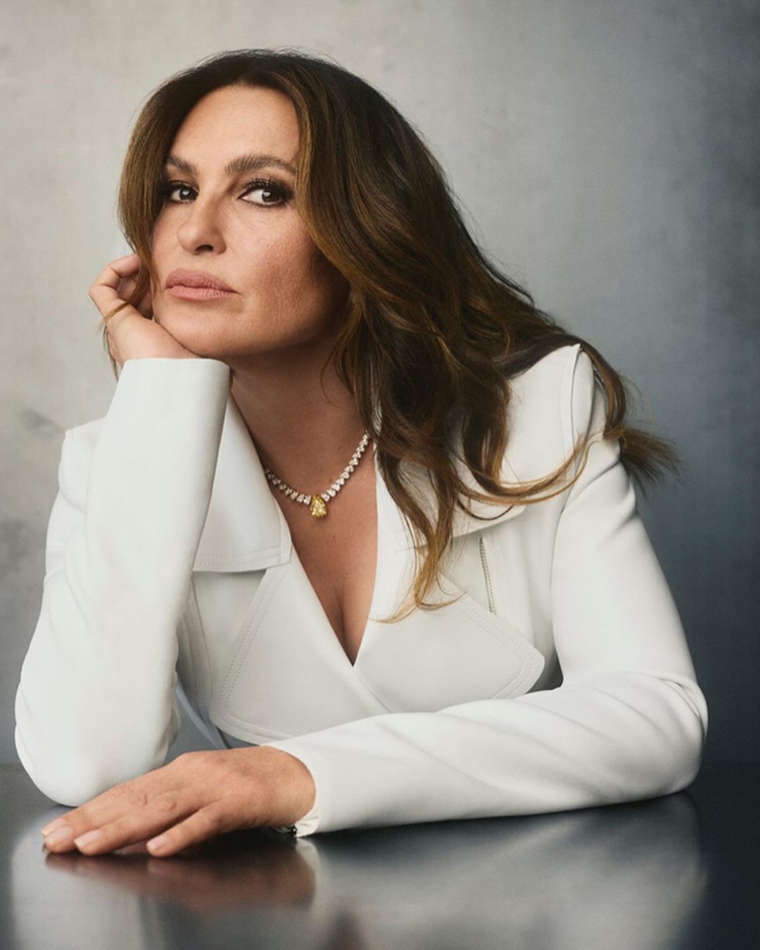 Portrait of a mature woman with brown hair wearing a white jacket and a yellow gemstone necklace at a reflective table.