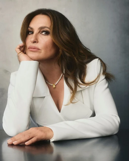 Portrait of a mature woman with brown hair wearing a white jacket and a yellow gemstone necklace at a reflective table.