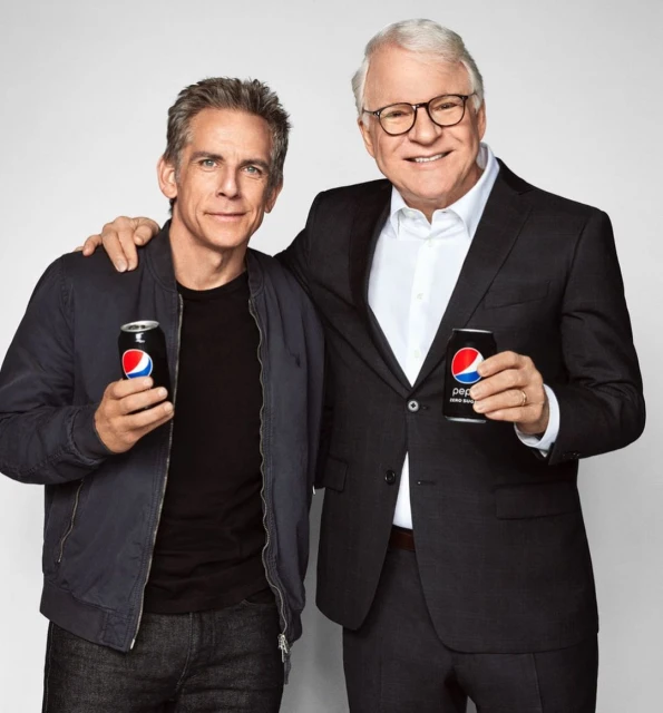 Two adult males, Steve Martin and Ben Stiller, pose together indoors holding Pepsi Zero cans against a plain backdrop in a medium shot.