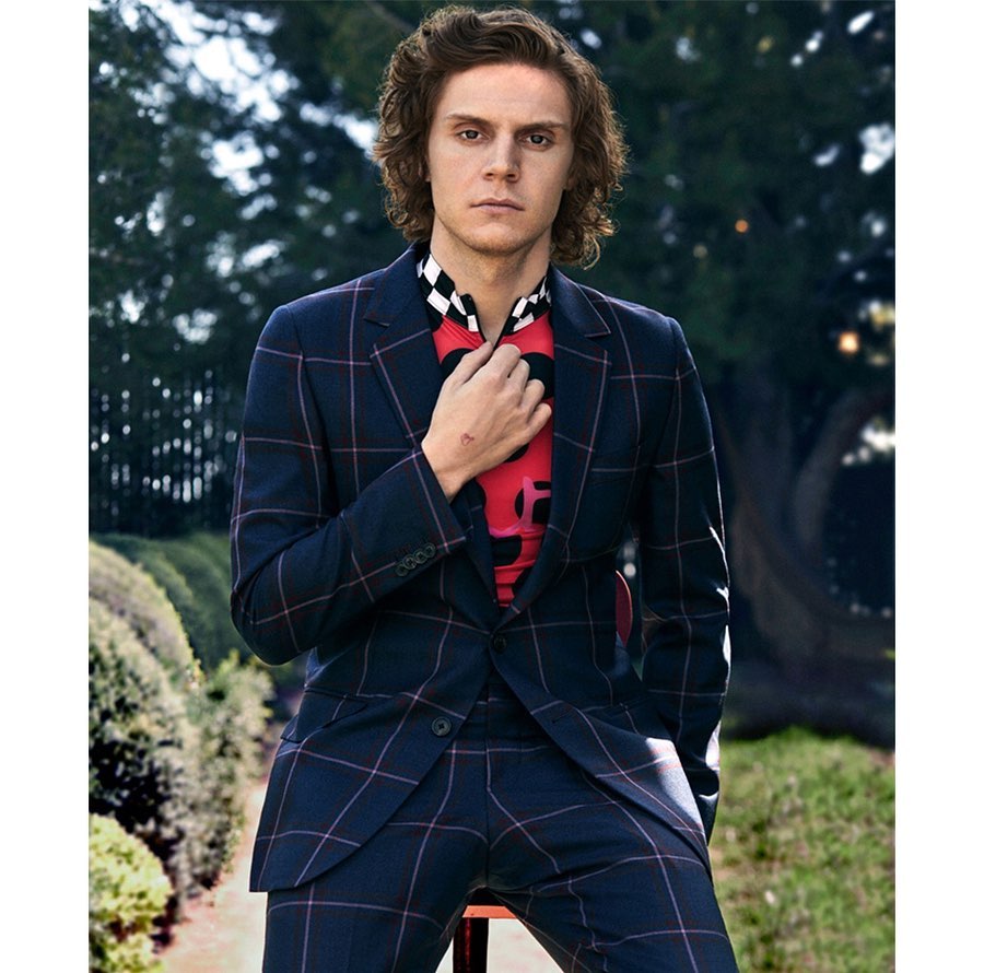 An adult male poses while seated outdoors, wearing a navy blue plaid suit with a colorful shirt and his hand at his neck.