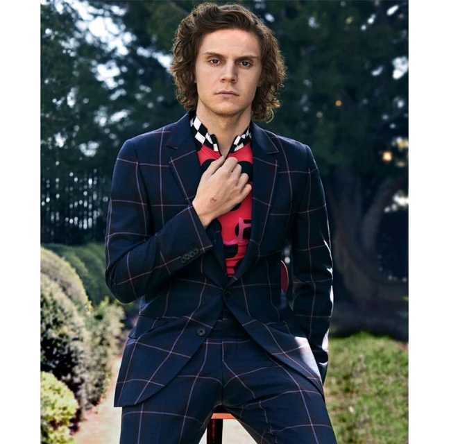 An adult male poses while seated outdoors, wearing a navy blue plaid suit with a colorful shirt and his hand at his neck.