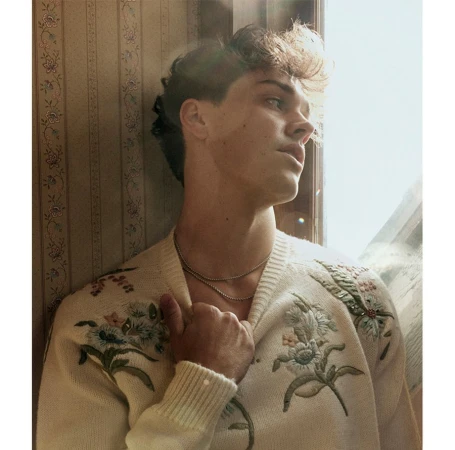 Young man wearing a cream embroidered cardigan standing against a patterned vintage wallpaper background