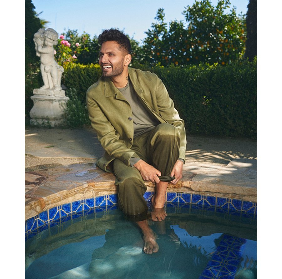 A man in an olive suit sits on the edge of a pool, feet in the water, in a garden setting with roses and greenery.