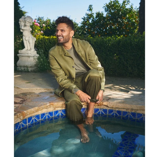 A man in an olive suit sits on the edge of a pool, feet in the water, in a garden setting with roses and greenery.