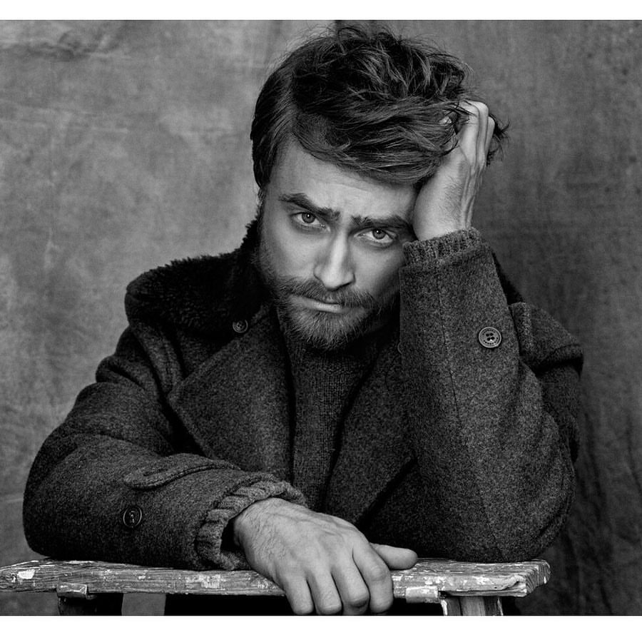Portrait of Daniel Radcliffe with a beard wearing a heavy wool coat, resting his hand in his hair against a textured backdrop.