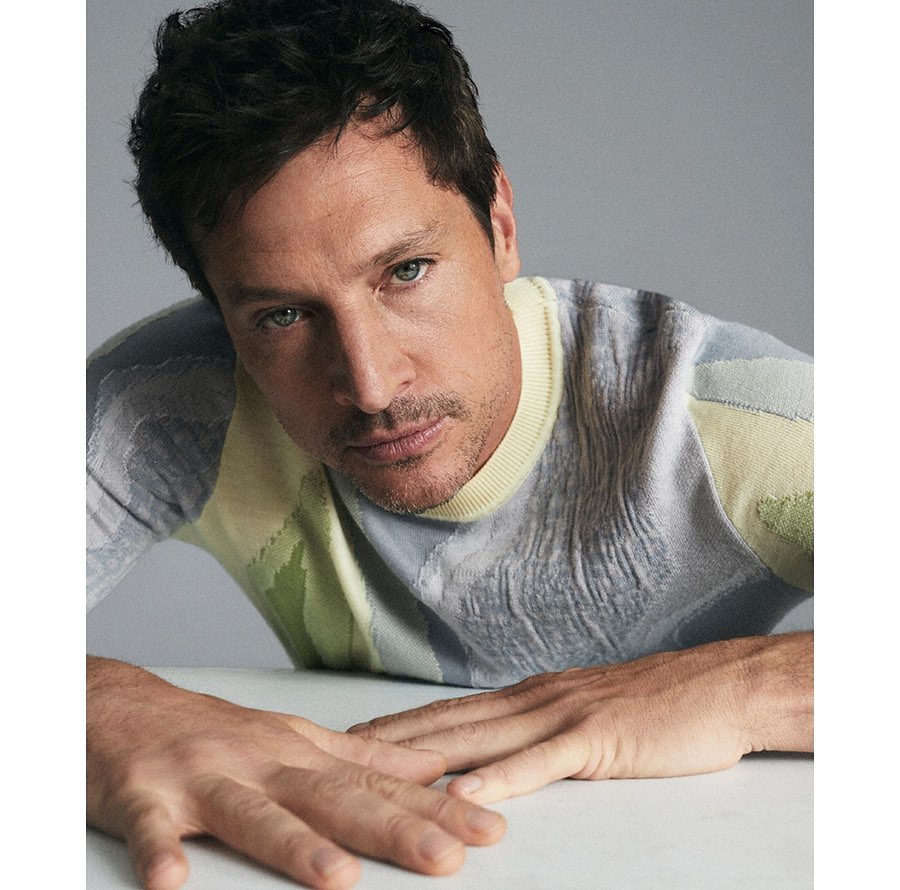 Portrait of a man wearing a patterned knit sweater leaning forward with hands resting on a white surface