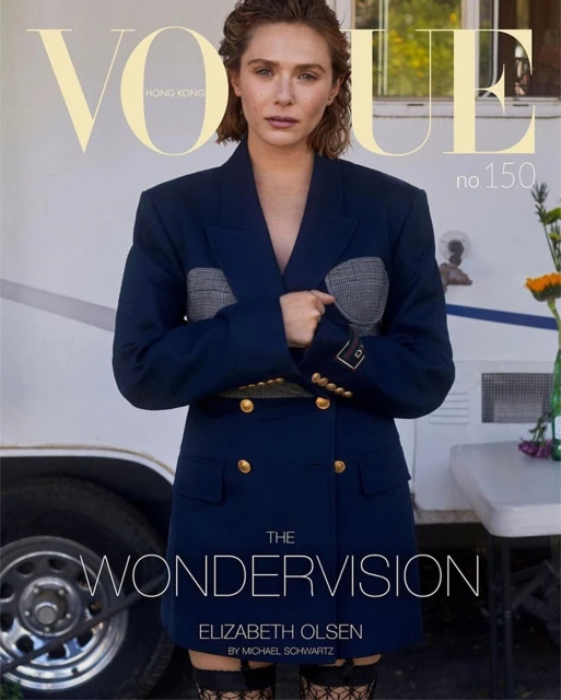 Elizabeth Olsen poses on the cover of Vogue Hong Kong with a trailer in the background, wearing a blue blazer dress and patterned hosiery.