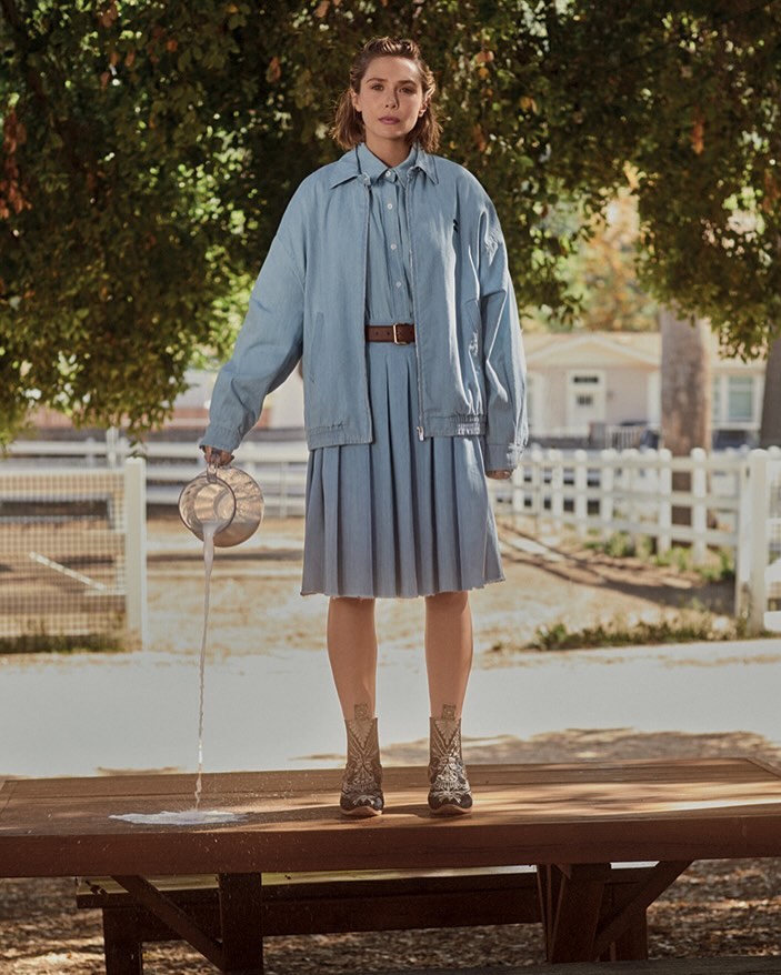A woman standing on a table outdoors, pouring milk from a pitcher. She is wearing a matching blue dress and jacket with cowboy boots.