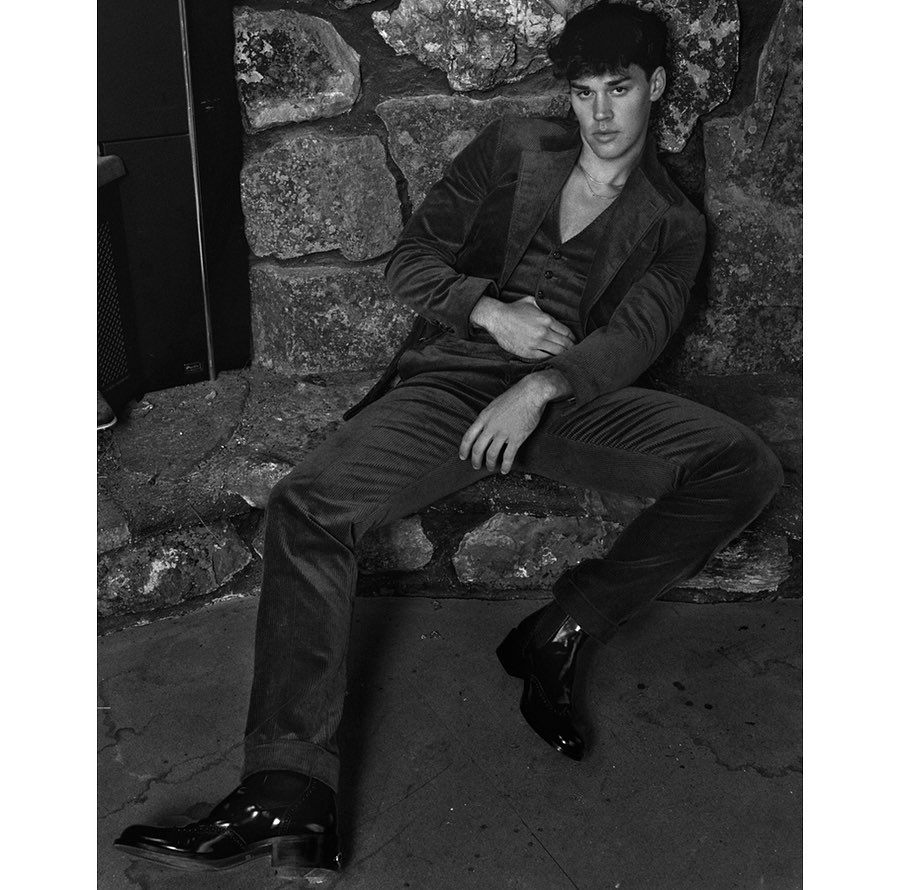 Monochrome portrait of a young adult male fashion model in a three-piece suit leaning against a stone wall, wearing dress shoes.