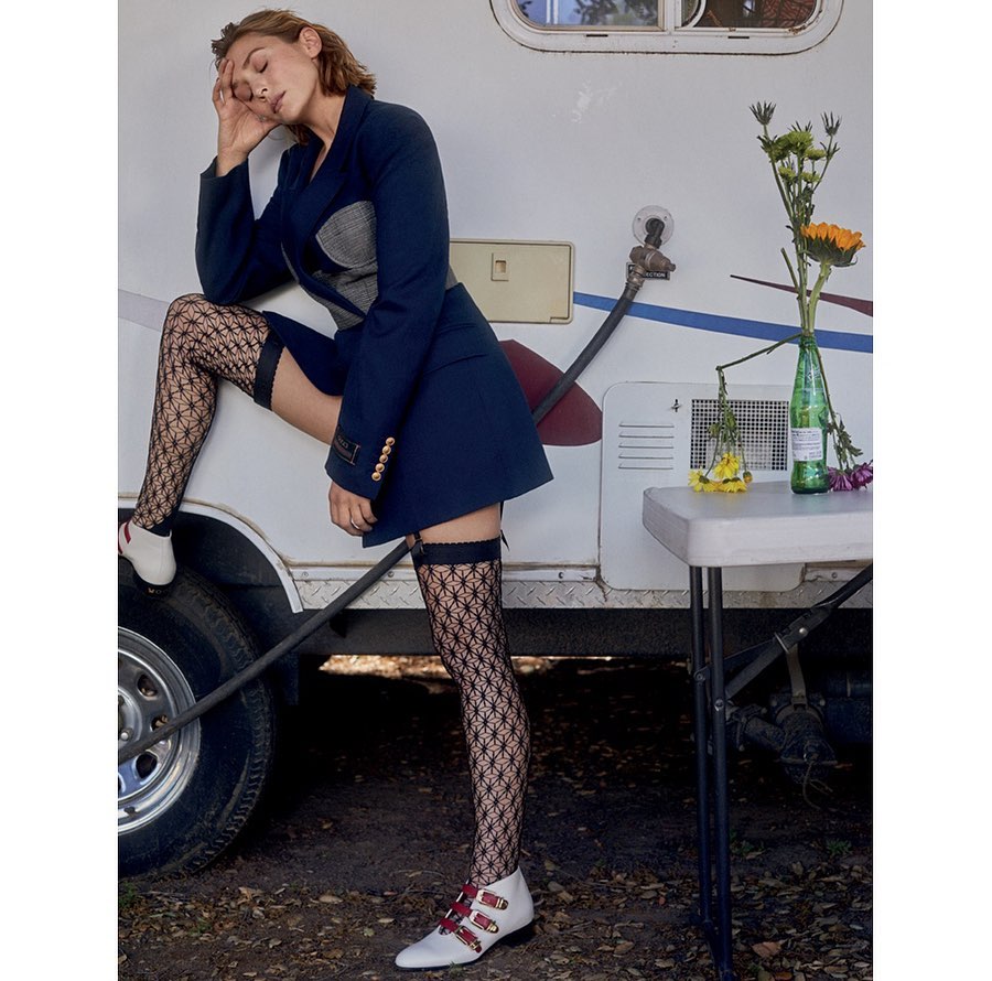 A woman poses outdoors wearing a short navy blazer dress, patterned thigh-high stockings, and white buckled shoes leaning against a trailer.