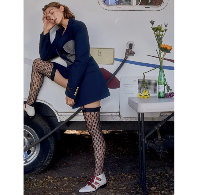 A woman poses outdoors wearing a short navy blazer dress, patterned thigh-high stockings, and white buckled shoes leaning against a trailer.