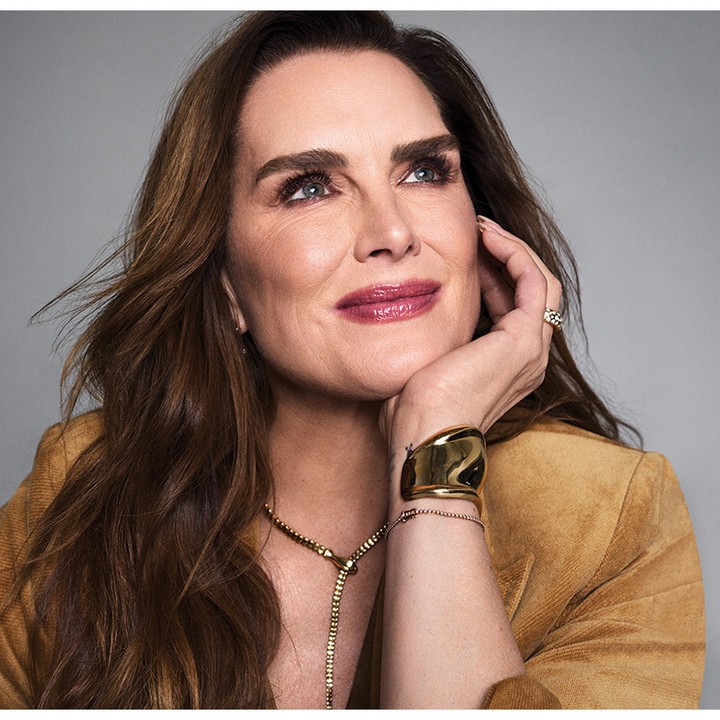 Close-up portrait of an adult woman in a tan jacket, featuring prominent jewelry, resting her head on her hand and looking upward.