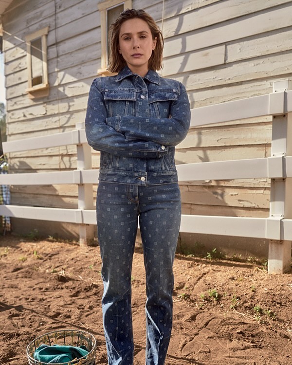 Elizabeth Olsen poses in denim jacket and jeans with a somber expression, set against a rustic outdoor backdrop with wooden fence and house.