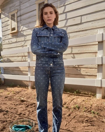 Elizabeth Olsen poses in denim jacket and jeans with a somber expression, set against a rustic outdoor backdrop with wooden fence and house.