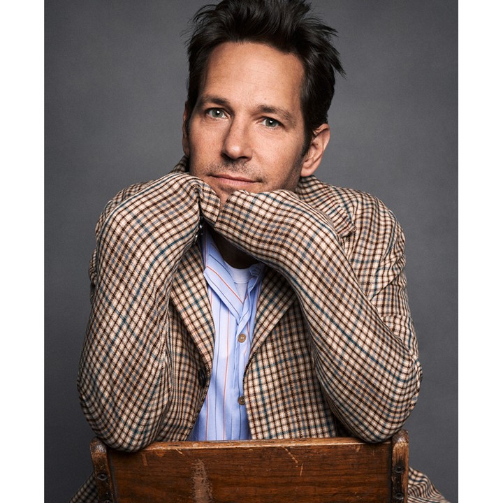 Portrait of actor Paul Rudd indoors against a grey backdrop. He is resting his chin on his hands while seated on a wood chair.
