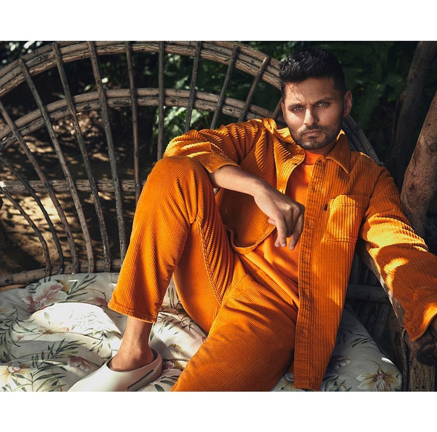 A stylish man sits in an outdoor chair in a corduroy orange outfit, set against a backdrop of green foliage and natural light.