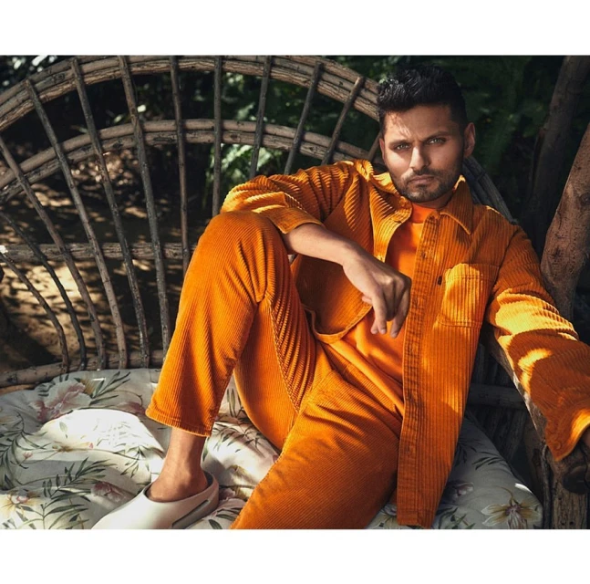 A stylish man sits in an outdoor chair in a corduroy orange outfit, set against a backdrop of green foliage and natural light.