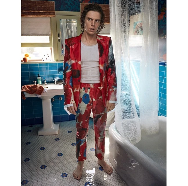 Man in a red floral suit stands barefoot in a bathroom with blue tile and a white bathtub, capturing a unique blend of fashion and interior.