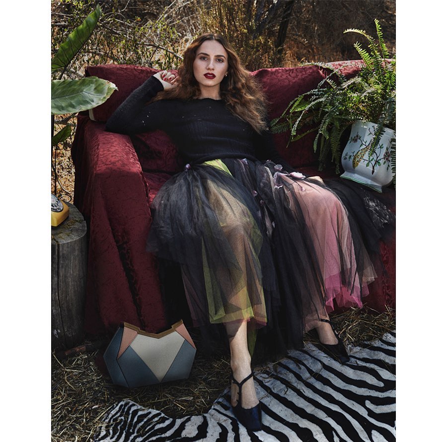 Woman in a black sweater and colorful tutu-style skirt poses on a red couch, accented with a zebra-striped rug and foliage backdrop.