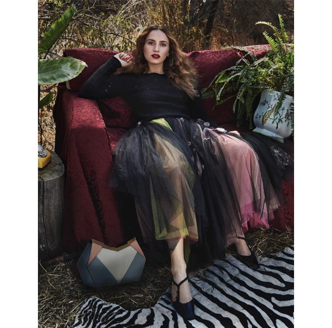 Woman in a black sweater and colorful tutu-style skirt poses on a red couch, accented with a zebra-striped rug and foliage backdrop.