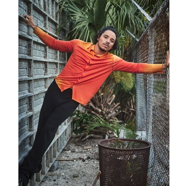 An adult male leans against a wall, wearing a brightly colored shirt and dark pants, with greenery and a wire fence in the background.