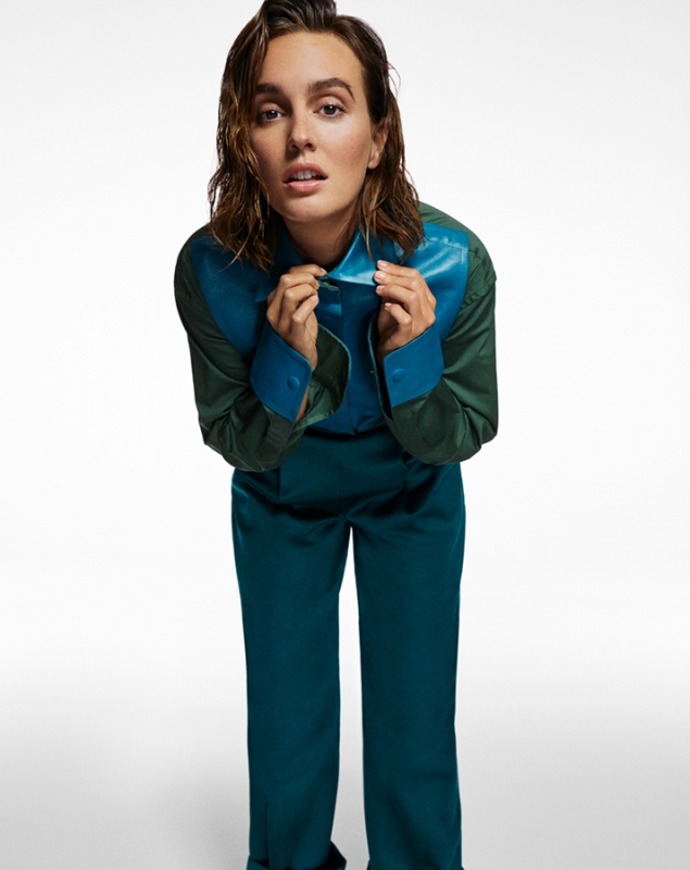 A fashionable adult female in a teal and green shirt and pants poses in a full shot against a white background with diffused lighting.