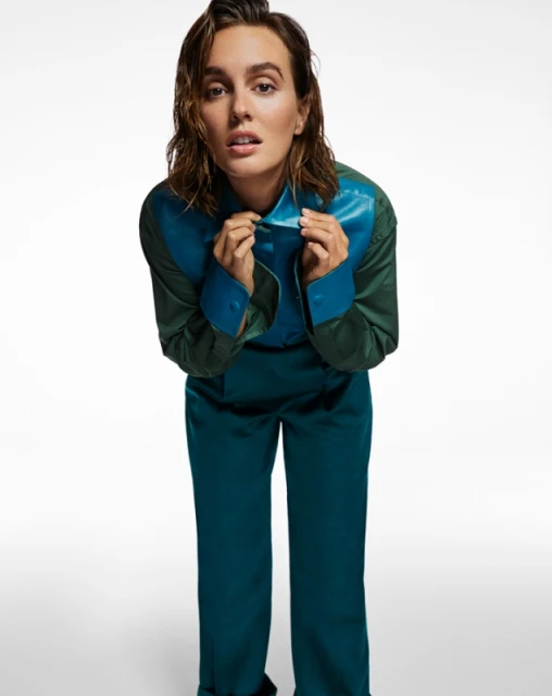 A fashionable adult female in a teal and green shirt and pants poses in a full shot against a white background with diffused lighting.