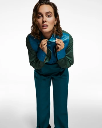 A fashionable adult female in a teal and green shirt and pants poses in a full shot against a white background with diffused lighting.