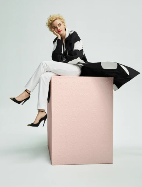 Woman with curly hair in patterned jacket and white pants poses seated on a pink cube in front of a light-grey background.
