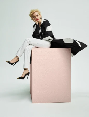 Woman with curly hair in patterned jacket and white pants poses seated on a pink cube in front of a light-grey background.