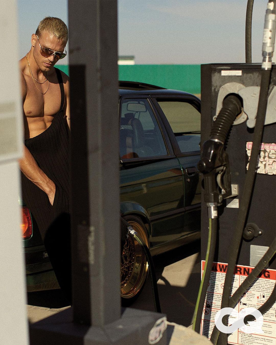 Adult male in sunglasses and black overalls stands next to a vintage car and gas pump at a gas station on a sunny day.