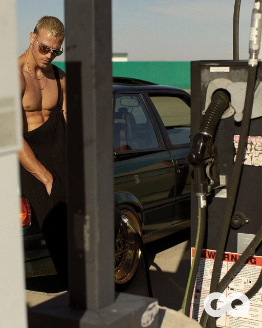 Adult male in sunglasses and black overalls stands next to a vintage car and gas pump at a gas station on a sunny day.