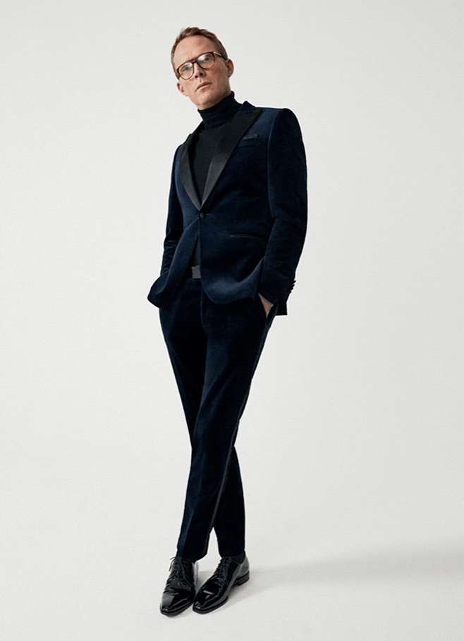 A man in a dark blue velvet suit and black turtleneck strikes a pose against a clean white background, showcasing contemporary fashion.