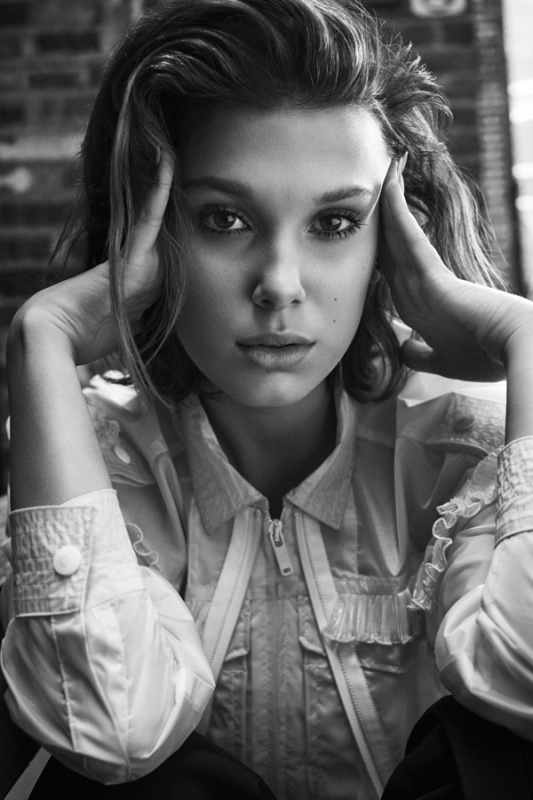 Portrait of a young woman with hands on her face, styled in an open collared shirt in black and white, brick wall background.