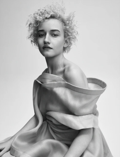 Portrait of Julia Garner in black and white, curly blonde hair, draped in light-colored fabric, in a studio setting, looking at the camera.